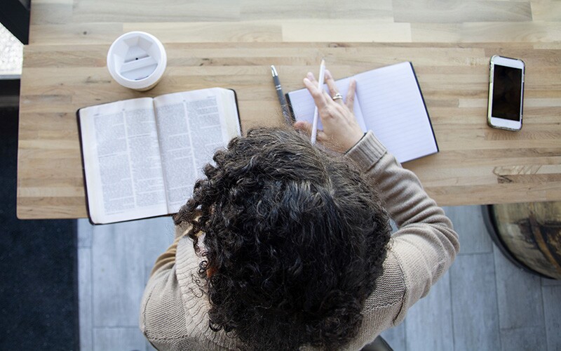 A woman studies scriptures with a scripture journal at her desk.