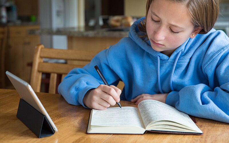 A young girl writes in her scripture journal as she studies the scriptures on a tablet.