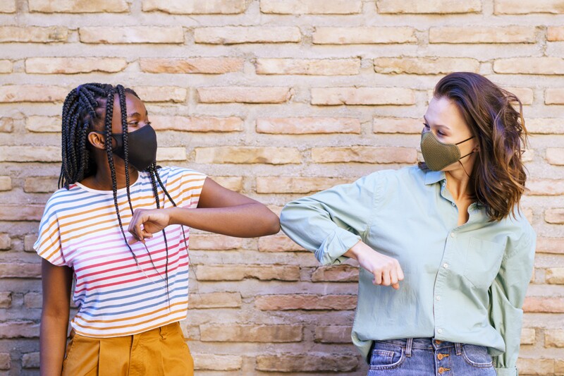two generation z girls wearing a mask and touching elbows.